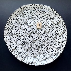 May include: A round decorative plate with a black rim, composed of white tiles with black polka dots. A wooden tile with the letter "T" is placed in the center of the plate. The plate is set against a black background.