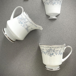 May include: A white porcelain tea set with a floral pattern in light blue and silver trim. The set includes a creamer, a teacup, and a decorative cup. The letter "T" is visible in the upper left corner.