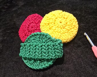 May include: Three circular, hand-crocheted scrubbers in bright yellow, magenta, and dark green. The green scrubber features an integrated strap across its center. A metal crochet hook with a pink handle is positioned to the right of the items.