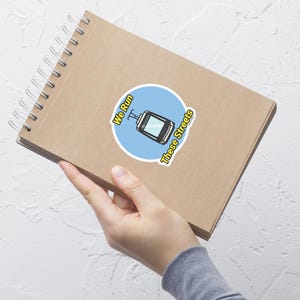 May include: A round sticker with a blue background featuring a cartoon cable car illustration. The sticker has the text "We Run These Streets" in a yellow font. The sticker is placed on a brown spiral notebook.