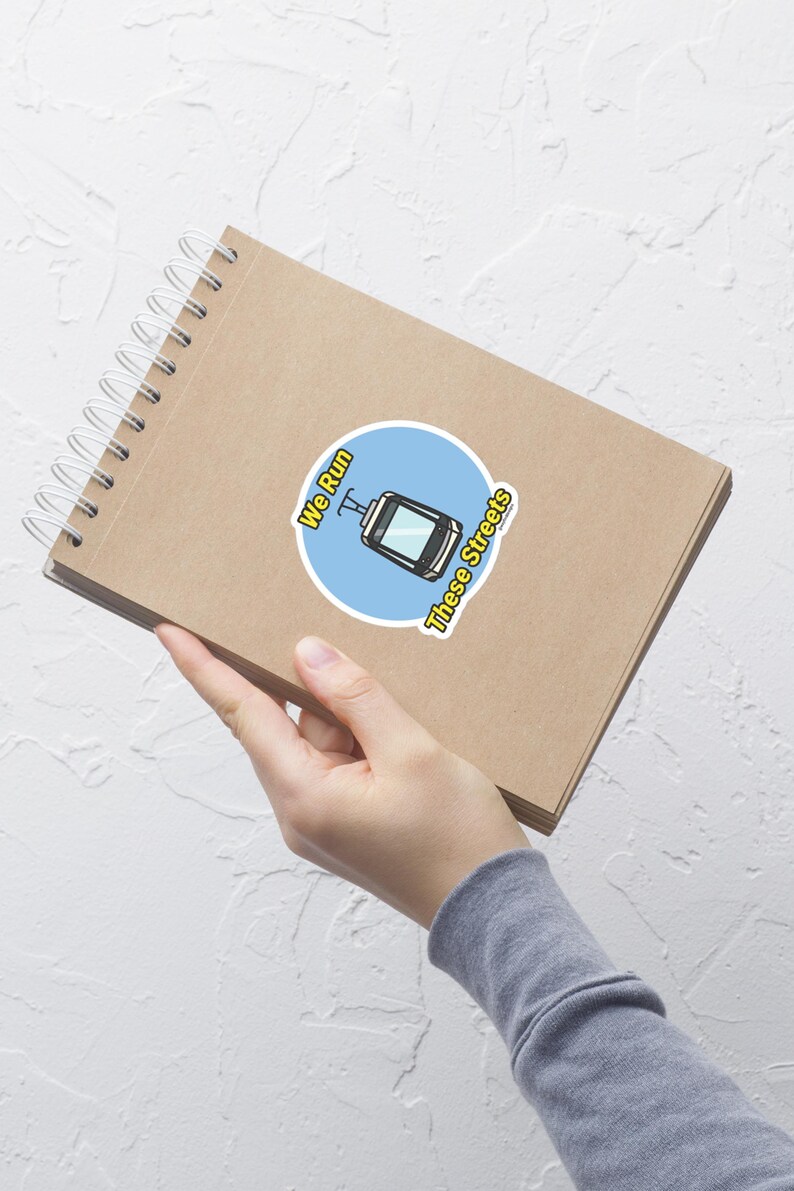 May include: A round sticker with a blue background featuring a cartoon cable car illustration. The sticker has the text "We Run These Streets" in a yellow font. The sticker is placed on a brown spiral notebook.