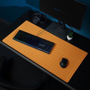 Pode incluir: Um teclado preto com iluminação arco-íris está sobre um grande tapete de rato com padrão laranja e castanho. Também há um rato de computador preto e uns auscultadores pretos sobre o tapete.