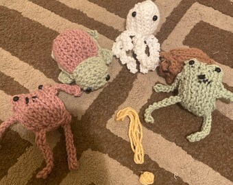 May include: Five crocheted stuffed animals, including a pink frog, a green frog, a pink and green turtle, a brown dog, and a white octopus. The animals are on a brown and white patterned rug. There is a yellow yarn question mark on the rug.