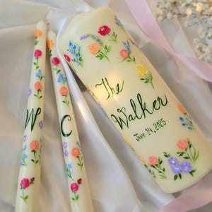 May include: A set of three off-white candles featuring hand-painted colorful floral patterns. The large pillar candle displays the text The Walker and June 14, 2025, while the two taper candles feature the individual letters W and C in green script.