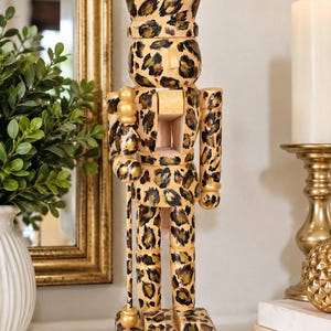 May include: A wooden nutcracker figure featuring a tan base colour with a black and dark brown leopard print pattern throughout. The figure wears a crown, holds a staff with spherical accents, and stands on a square base, all decorated in the same animal print design.