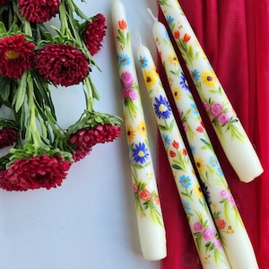 May include: Four white taper candles with hand-painted floral designs in shades of yellow, blue, red, and green. The candles are arranged on a red fabric background.