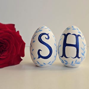 May include: Two white decorative Easter eggs with blue floral accents and the letters "S" and "H" painted in blue. A vibrant red rose is positioned to the left of the eggs.