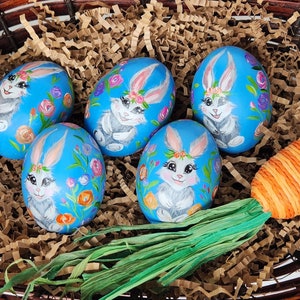 May include: A woven basket filled with decorative Easter eggs and carrots. The eggs are painted blue with a bunny and floral design. The carrots are orange with green tops, and the basket is lined with brown paper.