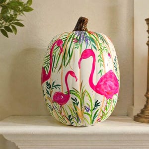 May include: A white decorative pumpkin featuring a hand-painted tropical design with vibrant pink flamingos, green palm leaves, and thin vertical gold lines. The pumpkin has a textured brown stem and a smooth, rounded, ribbed surface.