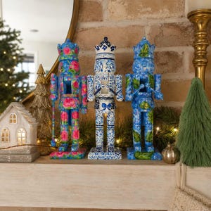 May include: Three decorative nutcracker figures standing in a row. The left figure features a pink and blue floral pattern. The center figure displays a blue and white botanical design. The right figure showcases a blue and green leaf motif.