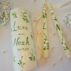 May include: A large pillar candle and two taper candles in off-white, hand-painted with green stems, leaves, and small white flowers. The pillar candle features the text "Luna and Noah" and "July 25, 2026". The taper candles feature the letters "L" and "N".