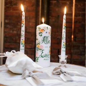 May include: A set of three white candles featuring hand-painted floral designs in yellow, pink, and green. The central pillar candle displays the text "Allie and Eric" with the date "09.12.25" in green script, while the two taper candles show dates.