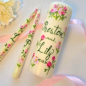 May include: Ivory-colored wedding candles with hand-painted pink flowers and green leaves. The large candle has the names "Preston and Lily" and the date "05.23.26". Two smaller candles have the letters "L" and "P".