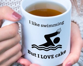 May include: White ceramic mug with black text and a graphic of a swimmer holding a cupcake. The text reads "I like swimming But I LOVE cake".