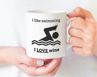 May include: White ceramic mug with a black silhouette of a person swimming and holding a glass of wine. The text "I like swimming I LOVE wine" is printed on the mug.