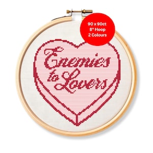 Op de afbeelding: Een kruissteekborduurwerk in een houten hoepel. Het ontwerp toont een roze hart met de woorden "Enemies to Lovers" in rood gestikt. De hoepel heeft een diameter van 20 cm en de stof is wit.