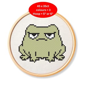 May include: Cross-stitch embroidery hoop featuring a grumpy green frog with black outlines. The text on the red oval reads: 45 x 35ct, colours = 3, Hoop = 5" or 6". The frog is centered on a white fabric, set in a wooden hoop.