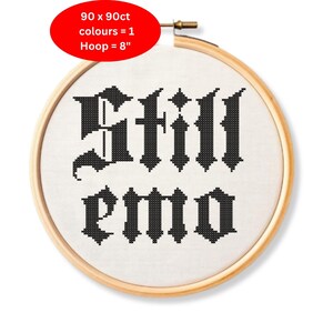 Puede incluir: Un bastidor de bordado de punto de cruz con las palabras "Still emo" en letras góticas negras. El bordado está sobre una tela blanca, y el bastidor tiene 20 cm de diámetro. Un óvalo rojo en la esquina superior izquierda indica "90 x 90ct colores = 1".