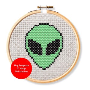 Puede incluir: Un bordado de punto de cruz de una cabeza alienígena en un aro de madera. El alienígena es verde con ojos negros y está bordado sobre una tela beige claro. El texto en el círculo rojo dice "Tiny Template 3" Hoop 504 stitches."
