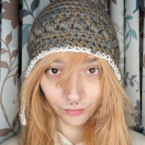 May include: A person wearing a brown and gray crocheted beanie with a white crocheted brim. The beanie has a long, thin string hanging from the side. The person has long, light brown hair and is wearing a white shirt.