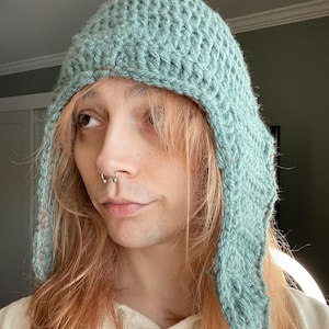 May include: A light blue crocheted hat with long ear flaps. The hat is made with a textured stitch and has a brown trim around the edges.