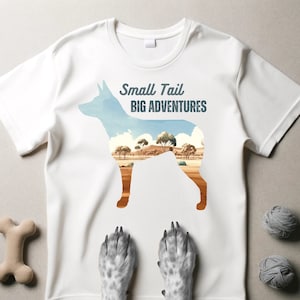 Può includere: Maglietta bianca con una silhouette di un cane con un paesaggio al suo interno. Il testo "Small Tail Big Adventures" è stampato sulla maglietta.