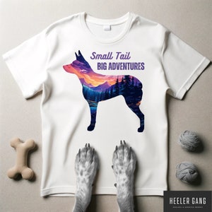 Può includere: Maglietta bianca con una silhouette di un cane con un paesaggio di tramonto al suo interno. Il testo "Small Tail Big Adventures" è stampato sulla maglietta.