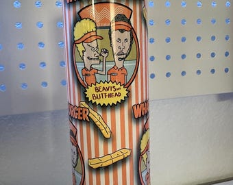 May include: A white tumbler with an orange and white striped background. The tumbler features a cartoon image of Beavis and Butt-Head in Whataburger uniforms. The text "Whataburger" is printed in orange on the top of the tumbler. The text "Beavis and Butt-Head" is printed in white on a yellow starburst. The tumbler also features a cartoon image of french fries.