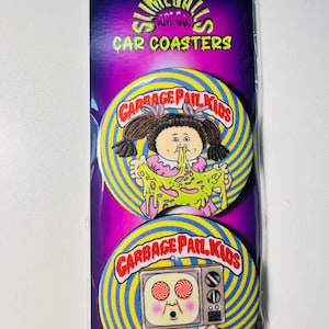 May include: Two car coasters featuring Garbage Pail Kids characters. The coasters are round with a yellow and blue spiral design. One coaster shows a girl with pigtails and green slime. The other shows a TV with red eyes.