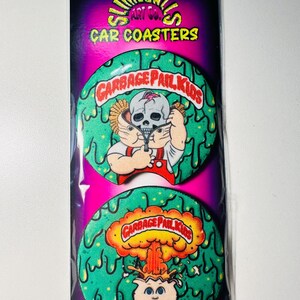 May include: Two car coasters featuring Garbage Pail Kids artwork. The coasters are green with a dripping design and feature cartoon characters. The top coaster shows a skull character, and the bottom coaster shows a character with an explosion.