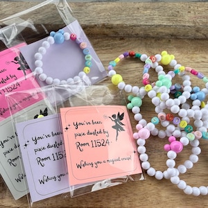 May include: White beaded bracelets with colorful accents and charms, some spelling out words. Packaged bracelets include cards with text and a fairy graphic. The text reads "You've been pixie dusted by Room 11524."