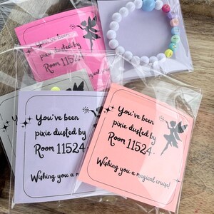 Może przedstawiać: Kolekcja małych prezent&oacute;w, w tym biała bransoletka z koralik&oacute;w z kolorowymi koralikami literowymi z napisem "WISH" oraz karty z napisem "You've been pixie dusted by Room 11524. Wishing you a magical cruise!" Prezenty są zapakowane w przezroczysty plastik.