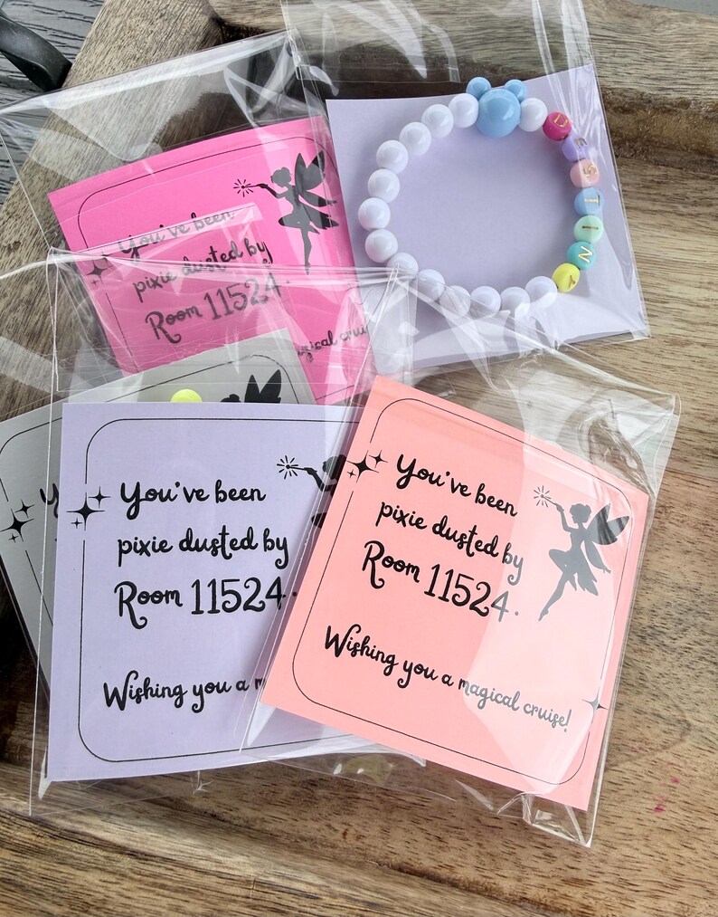 Może przedstawiać: Kolekcja małych prezent&oacute;w, w tym biała bransoletka z koralik&oacute;w z kolorowymi koralikami literowymi z napisem "WISH" oraz karty z napisem "You've been pixie dusted by Room 11524. Wishing you a magical cruise!" Prezenty są zapakowane w przezroczysty plastik.