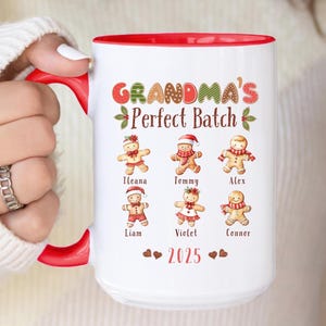 May include: A white ceramic mug with a red interior and handle, featuring the text "GRANDMA'S Perfect Batch" and gingerbread people with names. The year "2025" is also printed on the mug. The mug is held by a person.