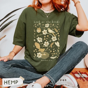 May include: An olive green t-shirt featuring a graphic print with the text "Look at the birds" and "Matthew 6:26". The design includes illustrations of birds, a nest with eggs, butterflies, feathers, and various flowers in shades of cream and gold.