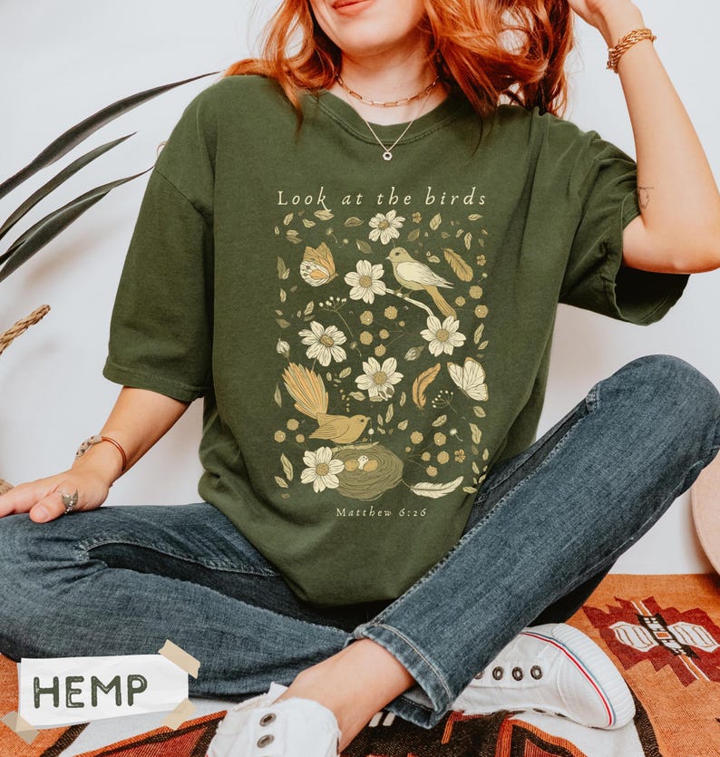 May include: An olive green t-shirt featuring a graphic print with the text "Look at the birds" and "Matthew 6:26". The design includes illustrations of birds, a nest with eggs, butterflies, feathers, and various flowers in shades of cream and gold.