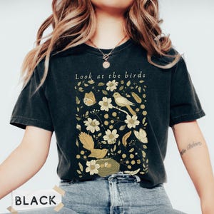 May include: A black short-sleeve t-shirt featuring a graphic print of birds, a nest, butterflies, and flowers in shades of cream, yellow, and olive green. The text "Look at the birds" is printed in white above the botanical and avian illustration.