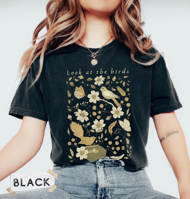 May include: A black short-sleeve t-shirt featuring a graphic print of birds, a nest, butterflies, and flowers in shades of cream, yellow, and olive green. The text "Look at the birds" is printed in white above the botanical and avian illustration.