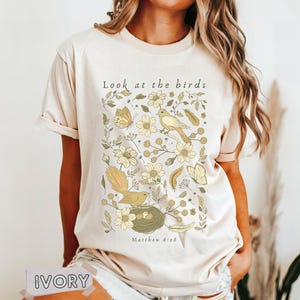 May include: An ivory short-sleeve t-shirt featuring a graphic print of birds, butterflies, flowers, and feathers in shades of yellow and green. The text "Look at the birds" is printed above the illustration, and "Matthew 6:26" is printed below.