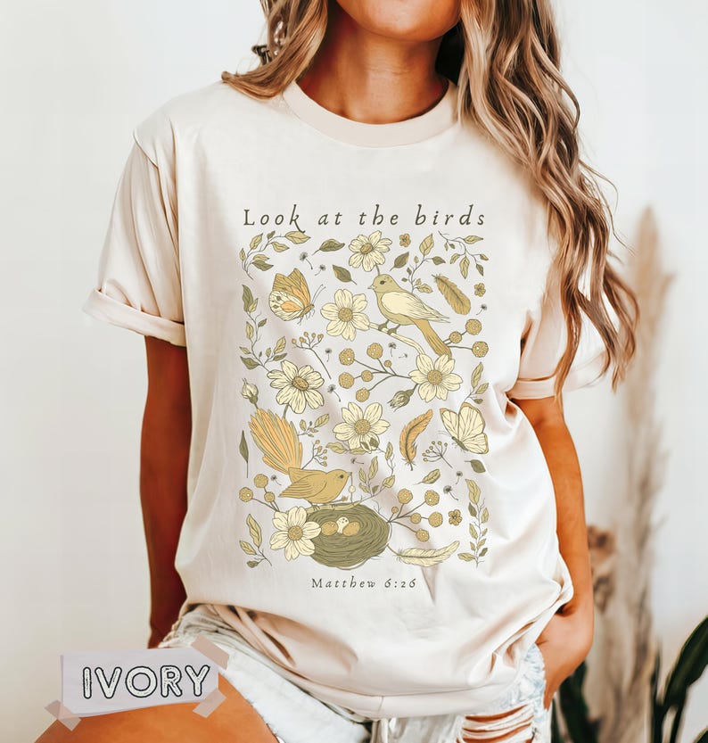 May include: An ivory short-sleeve t-shirt featuring a graphic print of birds, butterflies, flowers, and feathers in shades of yellow and green. The text "Look at the birds" is printed above the illustration, and "Matthew 6:26" is printed below.
