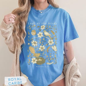 May include: A light blue t-shirt featuring a graphic print of birds, butterflies, flowers, and feathers in shades of yellow and brown. The text "Look at the birds" is printed at the top, and "Matthew 6:26" is printed at the bottom of the design.