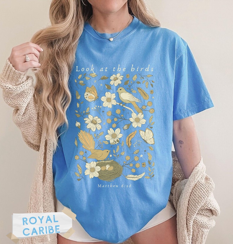 May include: A light blue t-shirt featuring a graphic print of birds, butterflies, flowers, and feathers in shades of yellow and brown. The text "Look at the birds" is printed at the top, and "Matthew 6:26" is printed at the bottom of the design.