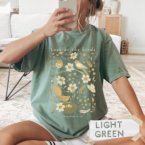 May include: A sage green short-sleeve t-shirt featuring a graphic print of birds, flowers, and butterflies in shades of yellow and cream. The text "Look at the birds" is printed at the top, and "Matthew 6:26" is printed at the bottom of the design.