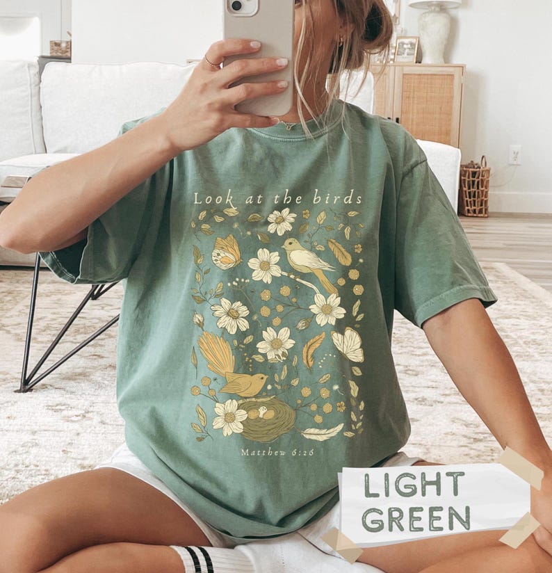 May include: A sage green short-sleeve t-shirt featuring a graphic print of birds, flowers, and butterflies in shades of yellow and cream. The text "Look at the birds" is printed at the top, and "Matthew 6:26" is printed at the bottom of the design.