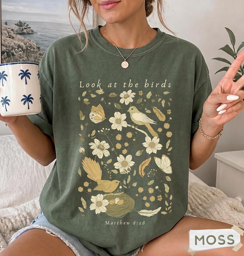 May include: An olive green t-shirt featuring a graphic print of birds, butterflies, flowers, and a nest. The text "Look at the birds" is printed above the illustration, and "Matthew 6:26" is printed below. The shirt has a crew neckline and short sleeves.