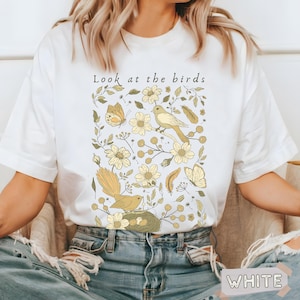 May include: A white short-sleeve t-shirt featuring a graphic print with the text "Look at the birds" above illustrations of yellow birds, butterflies, feathers, and various flowers. The design is centered on the chest of the garment.