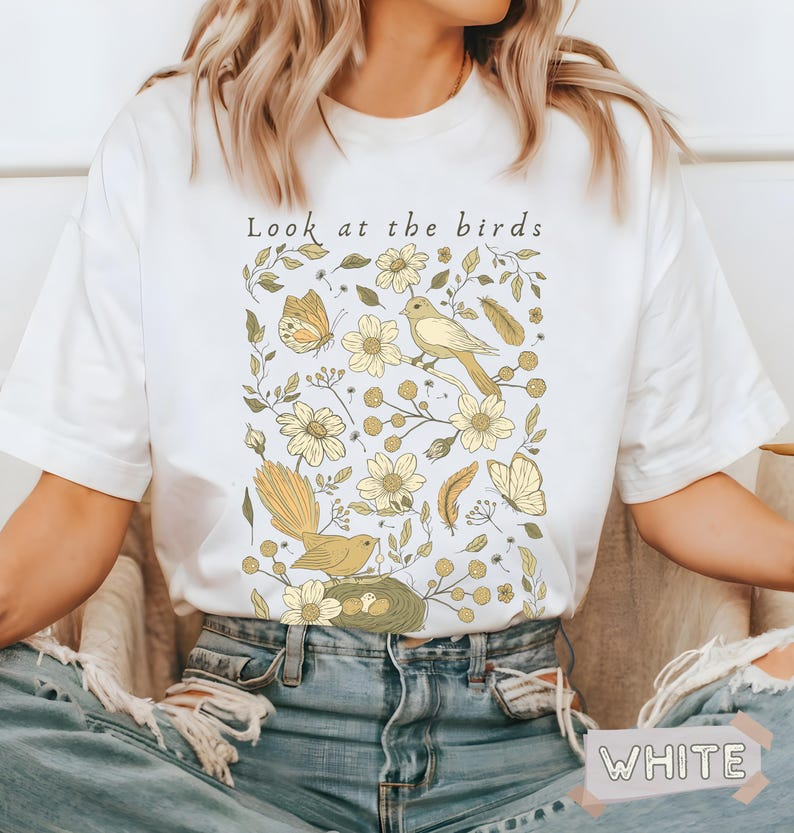 May include: A white short-sleeve t-shirt featuring a graphic print with the text "Look at the birds" above illustrations of yellow birds, butterflies, feathers, and various flowers. The design is centered on the chest of the garment.