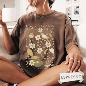 May include: A brown short-sleeve t-shirt featuring a graphic print of birds, butterflies, flowers, and a nest. The text "Look at the birds" is printed above the graphic, and "Matthew 6:26" is printed below. The shirt has a crew neckline.