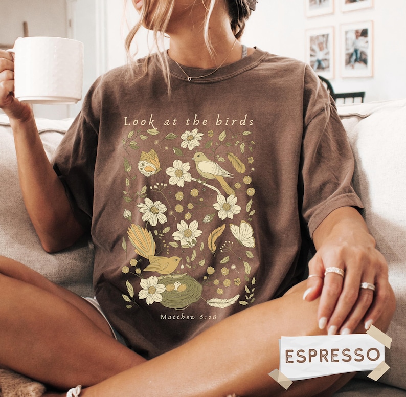 May include: A brown short-sleeve t-shirt featuring a graphic print of birds, butterflies, flowers, and a nest. The text "Look at the birds" is printed above the graphic, and "Matthew 6:26" is printed below. The shirt has a crew neckline.