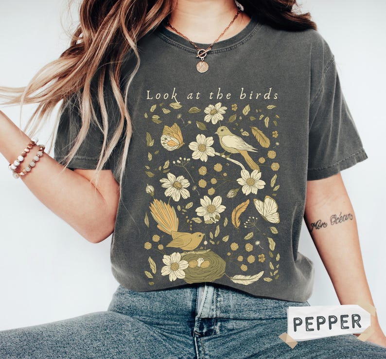 May include: Dark gray t-shirt with a nature-themed graphic. The design features birds, flowers, and butterflies in shades of yellow and cream, with the text "Look at the birds" at the top. The shirt is a casual style.
